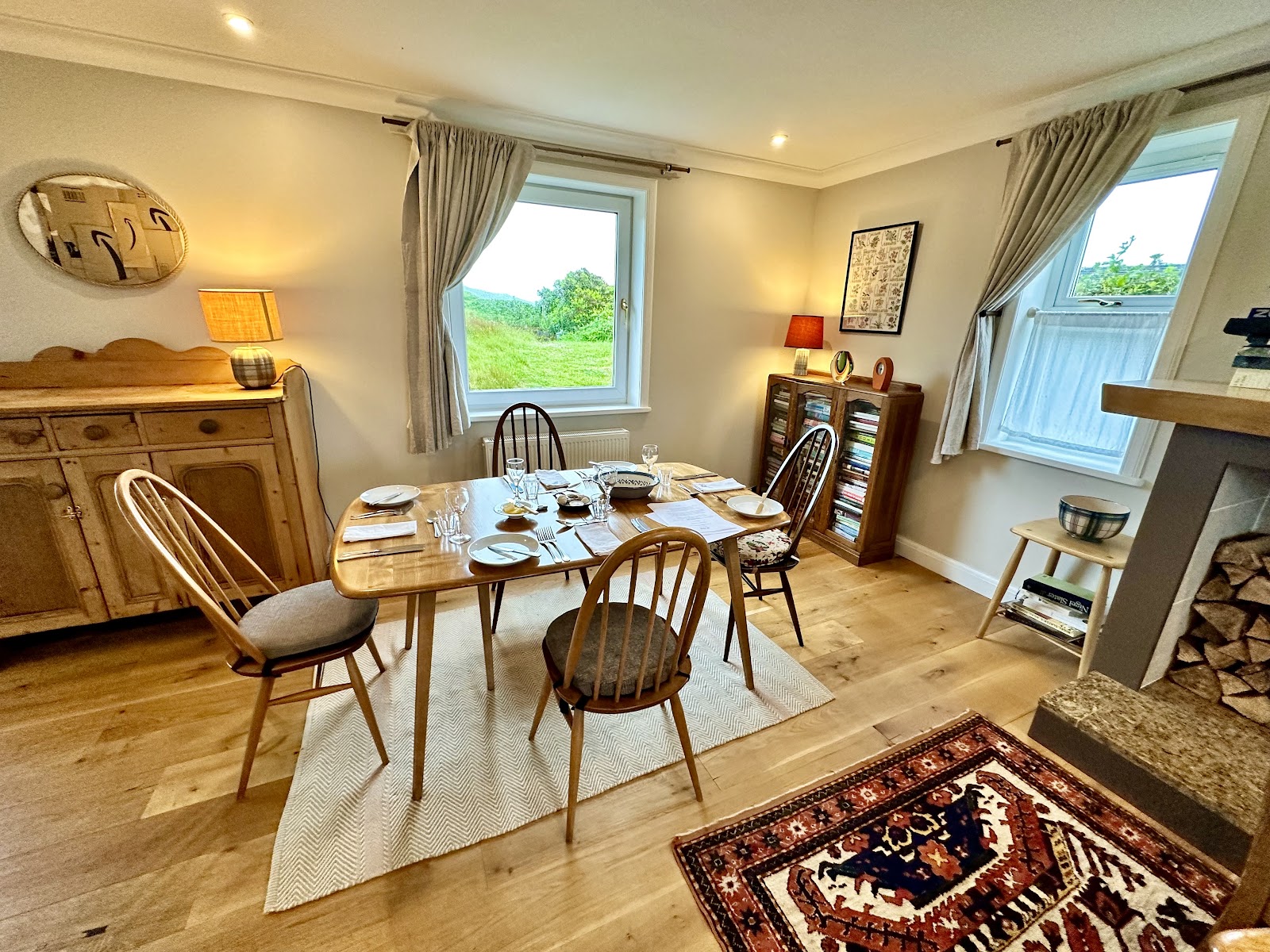The dining room at Ar Bòrd in Dervaig, a table laid for dinner on a herringbone rug, with a pine dresser, a bookcase and windows looking out on green fields.