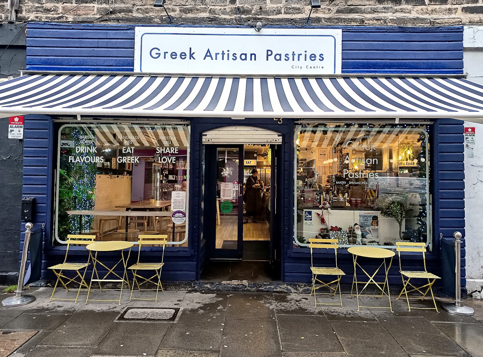 The navy front of Greek Artisan Pastries on Bread Street, under a striped awning, with yellow cafe tables and chairs outside on the pavement.