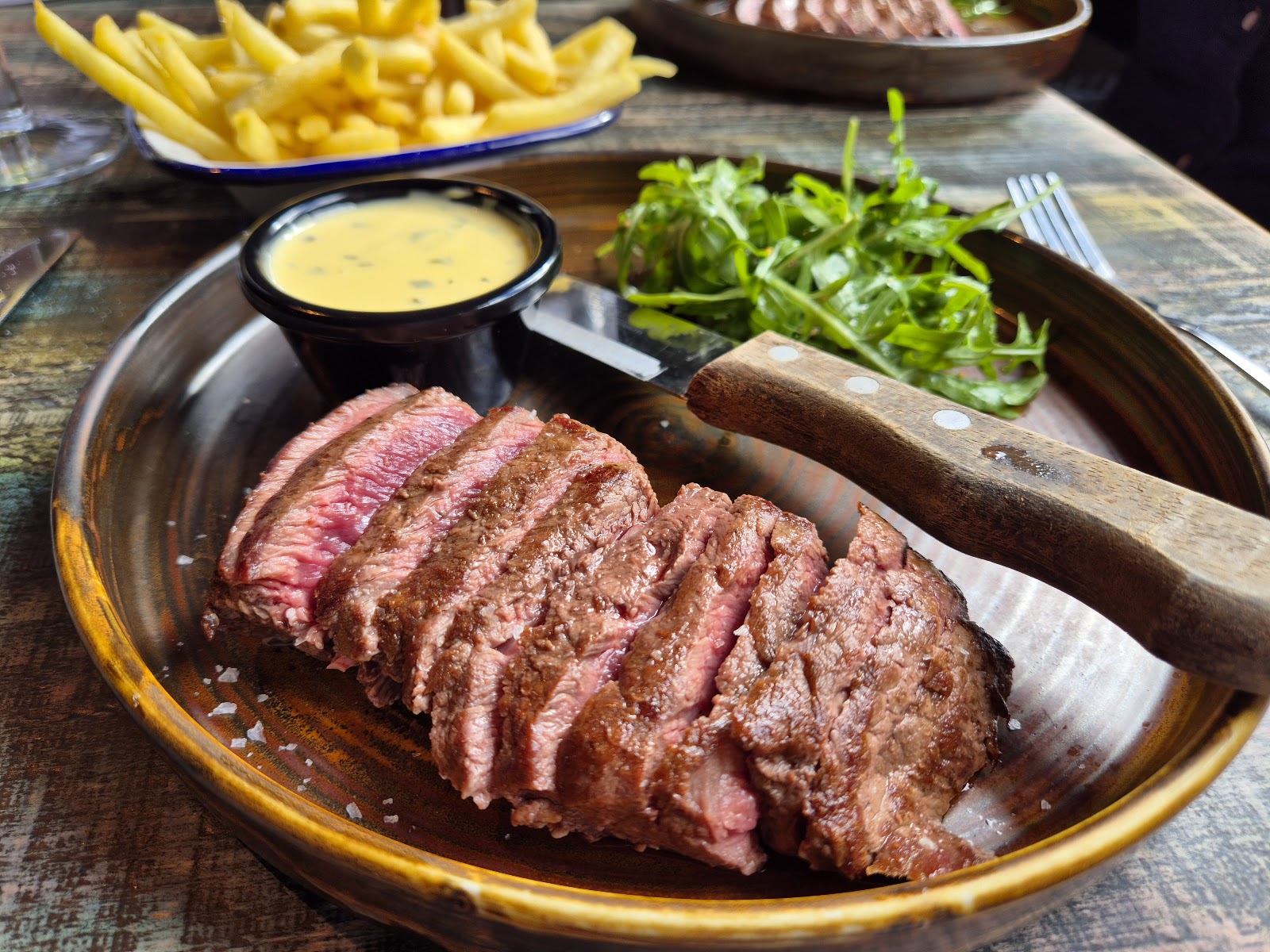 Sliced flatiron steak on a board with fries, salad and béarnaise.