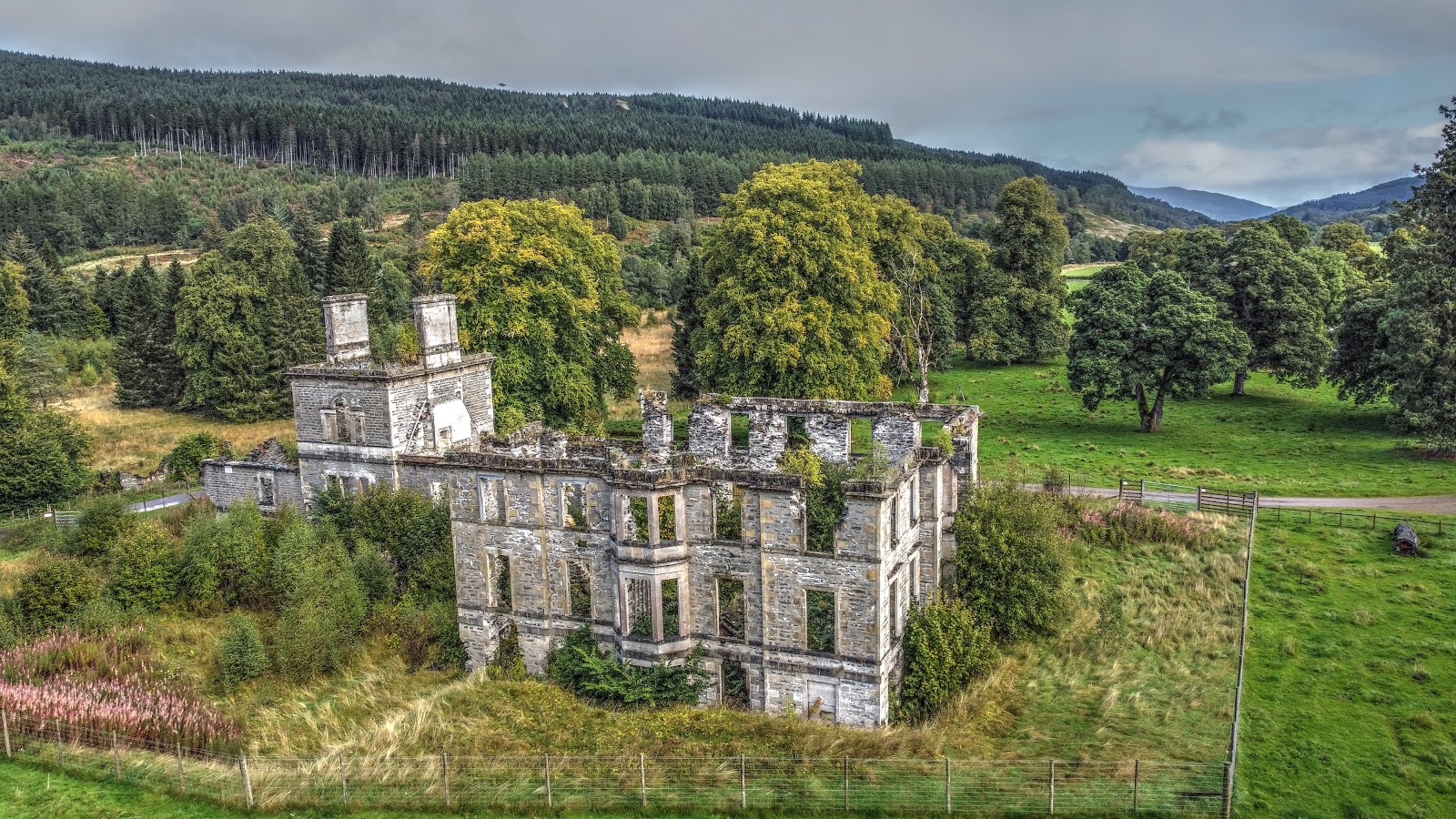 Guisachan House from the air: the roofless grey stone mansion with its two tall chimneys and empty windows, fenced off in long grass, with parkland trees and forested hills beyond.