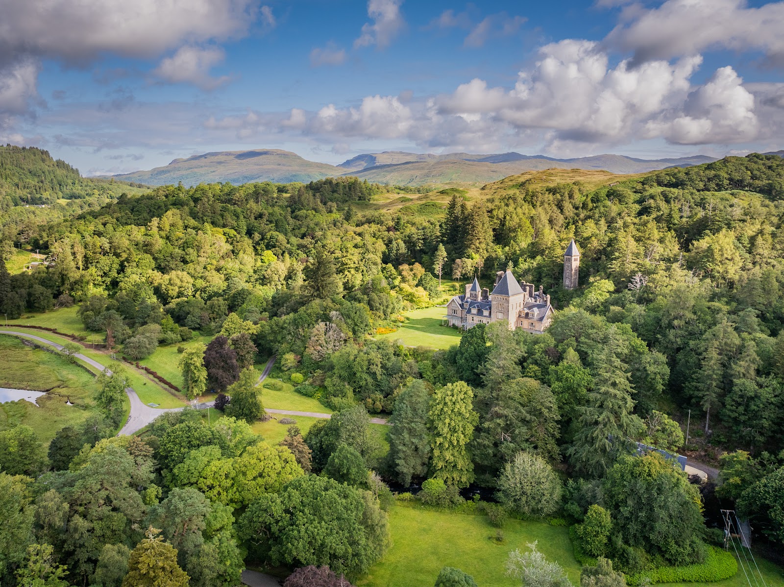 Ardtornish House in its gardens from the air, lawns and flowering shrubs round the pink sandstone mansion and its tower, woods all round and the Morvern hills beyond under summer cloud.