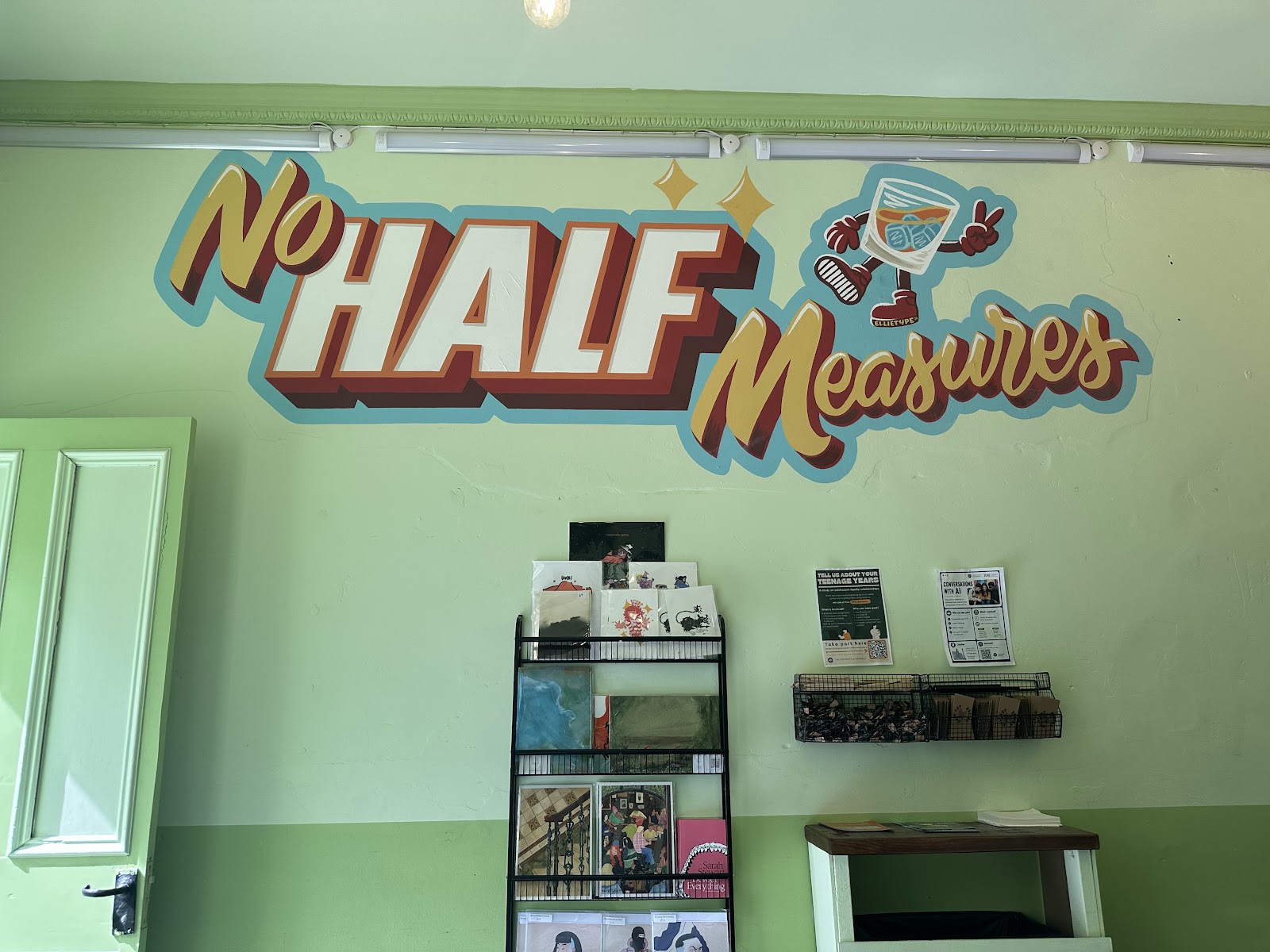 The painted 'No Half Measures' sign on the pale green wall inside Half-Cut Coffee, above a rack of prints and cards.