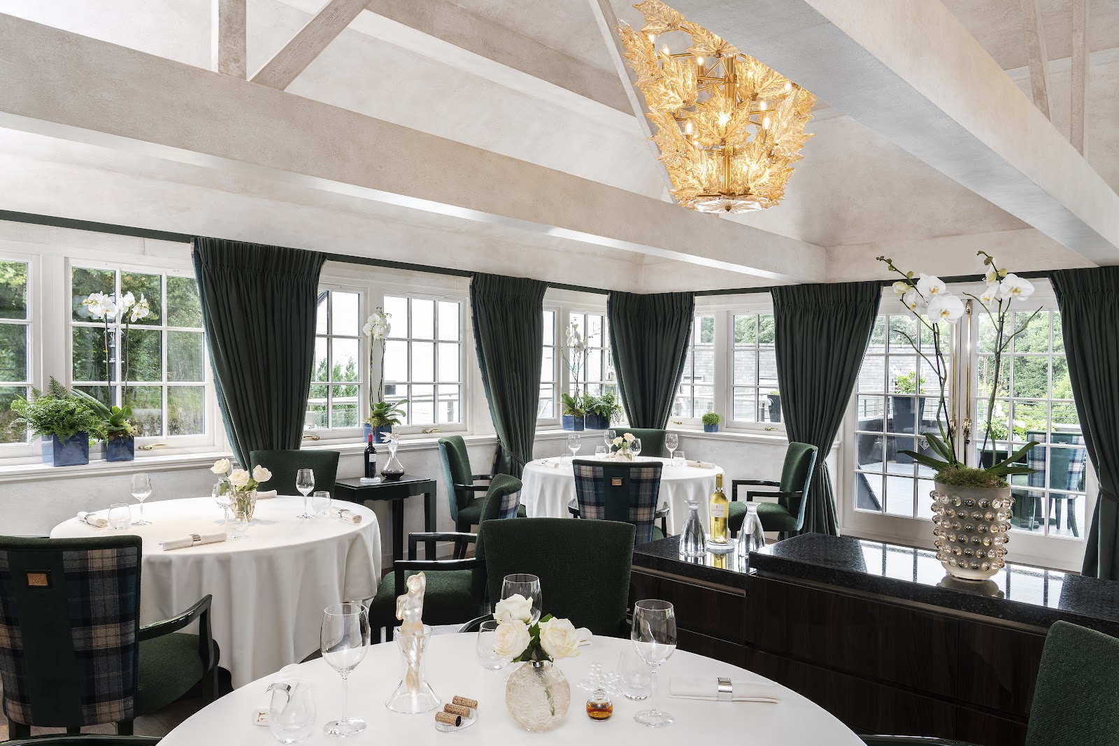 The dining room at the Glenturret Lalique Restaurant, with tables laid in white under a beamed ceiling, a golden Lalique chandelier, tartan chairs and tall windows onto the garden.