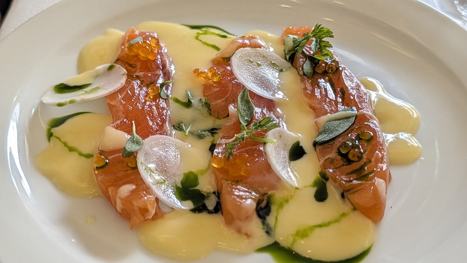 Slices of cured fish with a pale sauce, radish, herbs and roe on a white plate at eleanore.