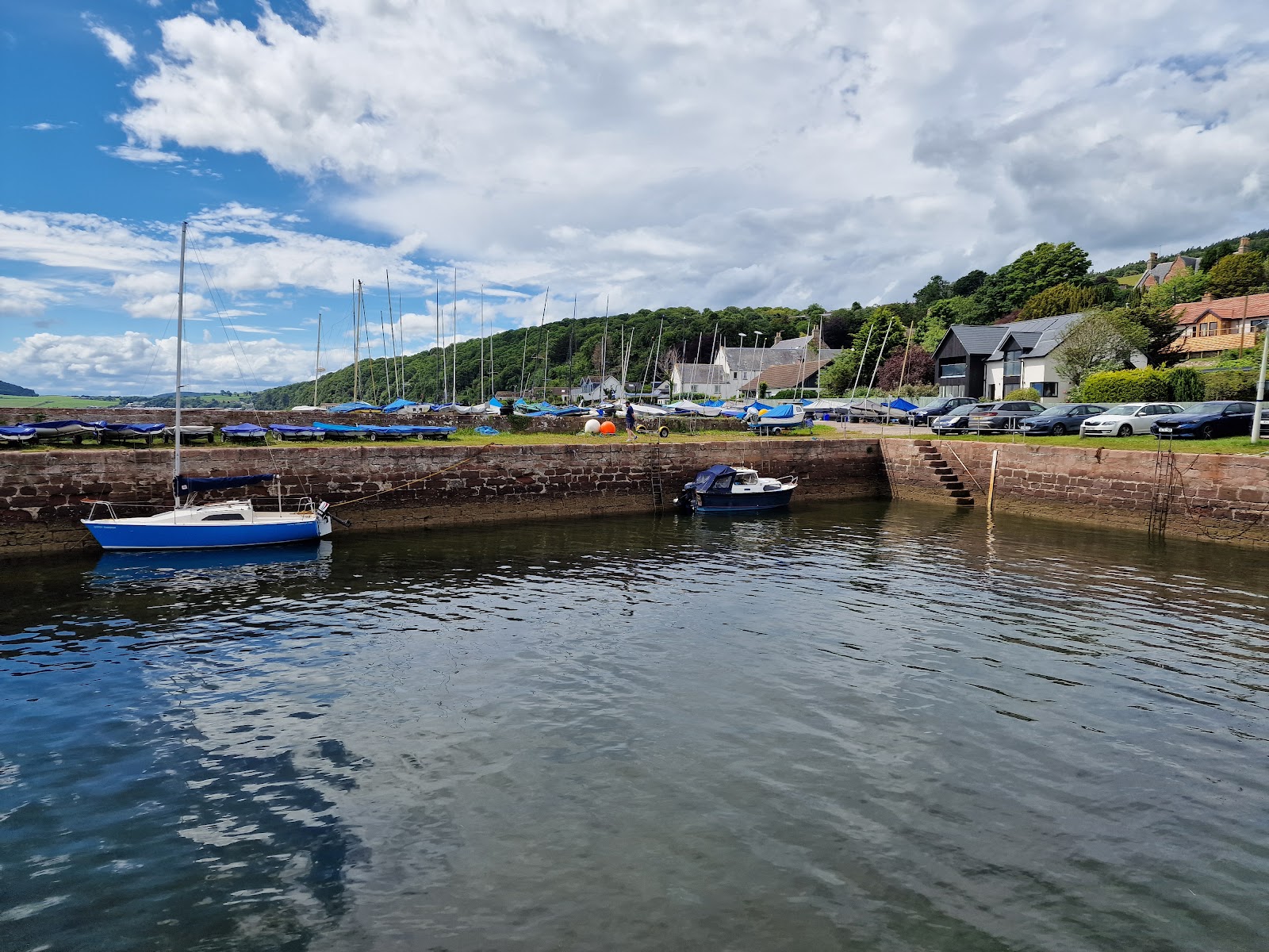 Small boats moored inside the sandstone walls of Fortrose Harbour on calm water, the town on green slopes behind.