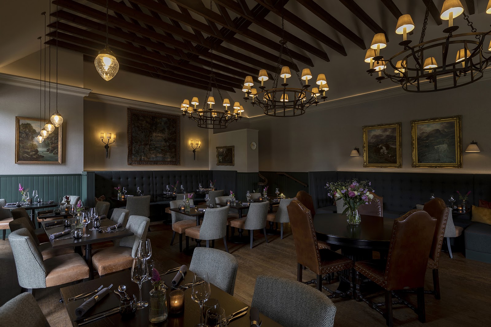 The dining room at Kildrummy Inn, with iron chandeliers under the beams, tables laid with glasses and candles and gilt-framed Highland paintings on the walls.