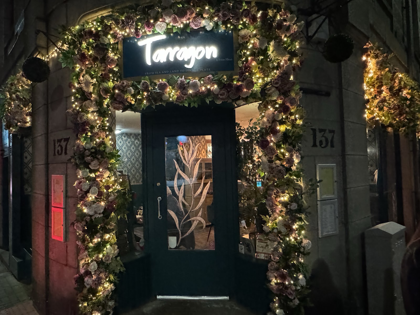 The door of Tarragon at 137 Rosemount Place at night, framed by an arch of artificial flowers and fairy lights under the lit Tarragon sign.