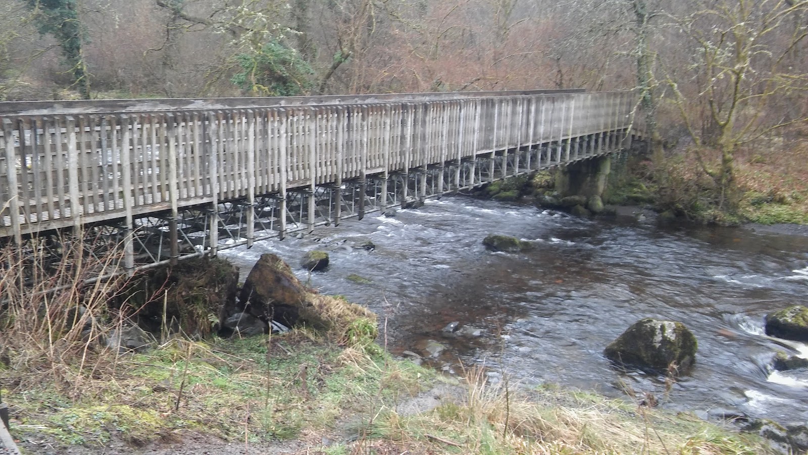 A long wooden footbridge on trestles crossing the River Carron, the water running fast over boulders below.