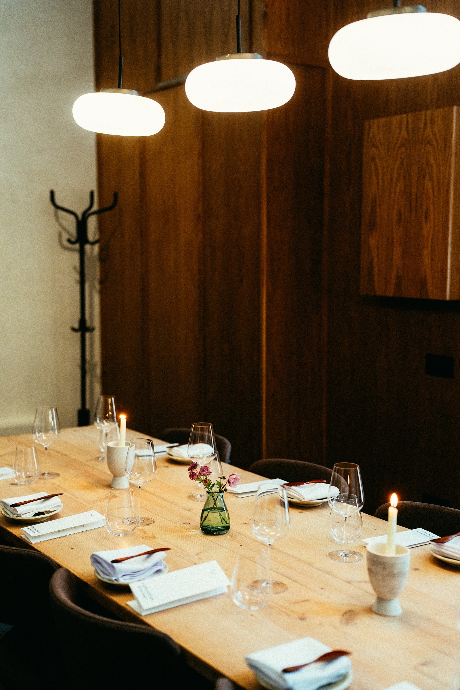 The long communal table at Fallachan Kitchen laid for dinner, with candles, flowers and pendant lights against wooden panelling.