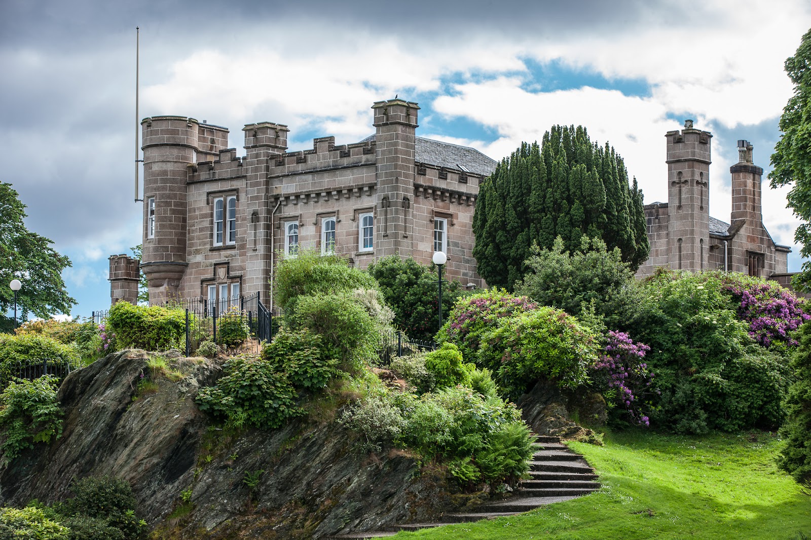 Castle House in Dunoon, a turreted sandstone villa with battlements, above a rocky bank of rhododendrons in flower, steps climbing the lawn beside it and the Castle Hill flagpole on the left.