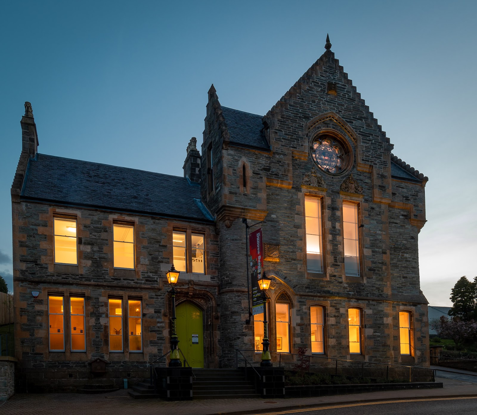 Dunoon Burgh Hall at dusk, a stepped-gable stone building with a round rose window, its tall windows lit and a green door between two street lamps.