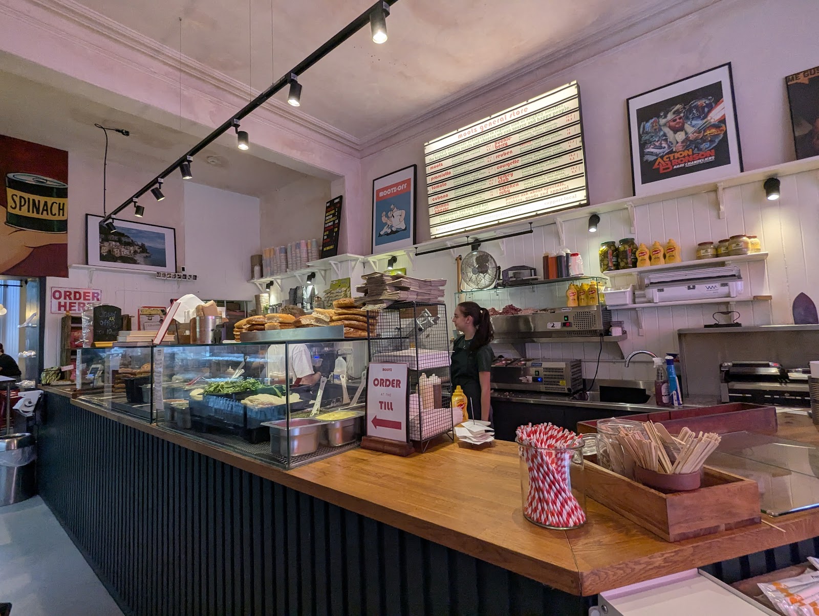 The counter at Mootz General Store on Raeburn Place, with a stack of schiacciata loaves, the sandwich board lit up on the wall and framed posters above the shelves.