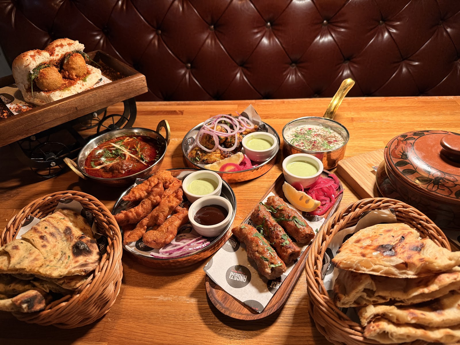 Seekh kebabs, fried chicken, a curry, chutneys and baskets of naan on a table at Angeethi.