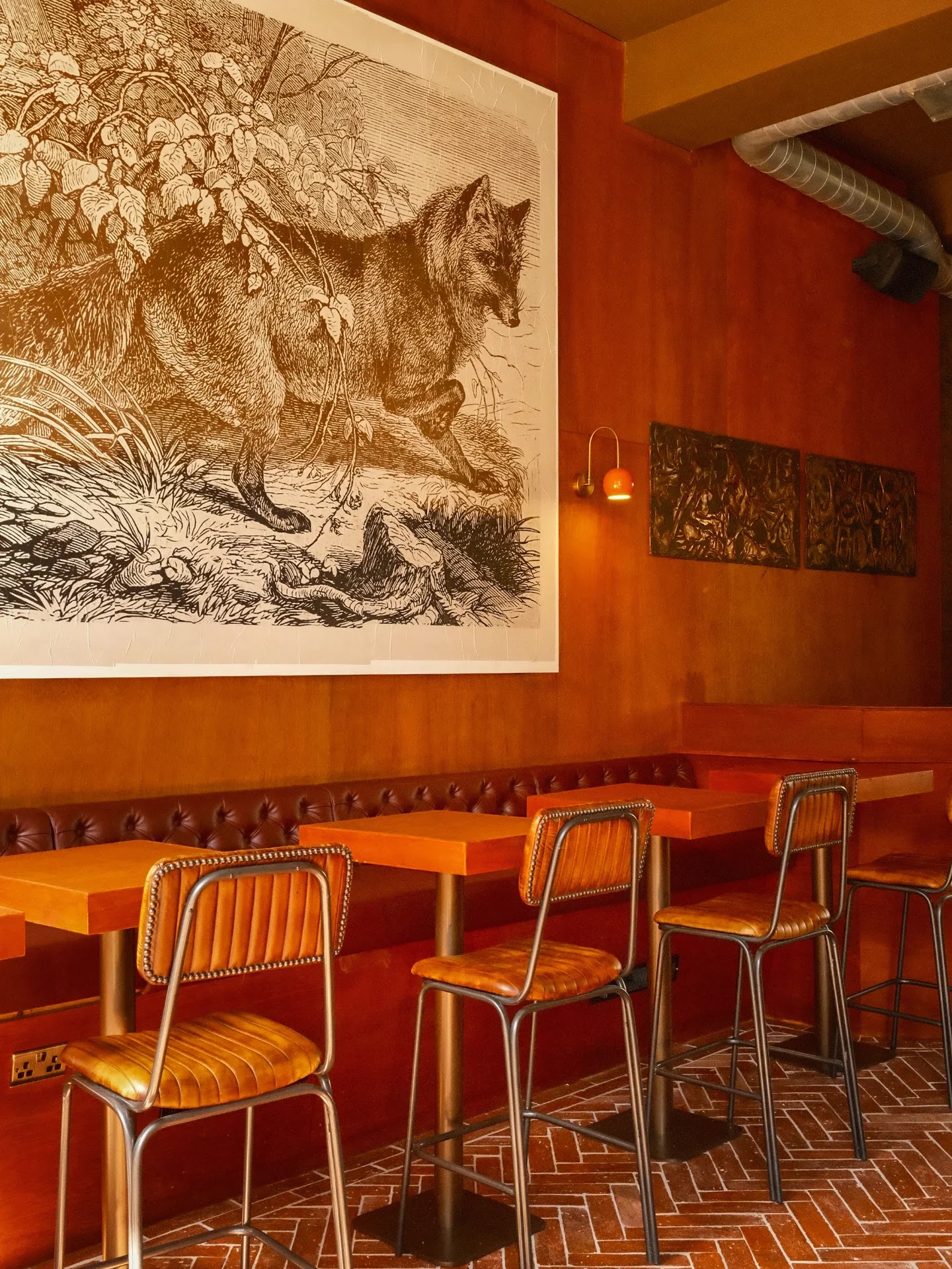 High stools and tables along a leather banquette at Bar Dani, under a large engraving of a fox on warm wooden walls.