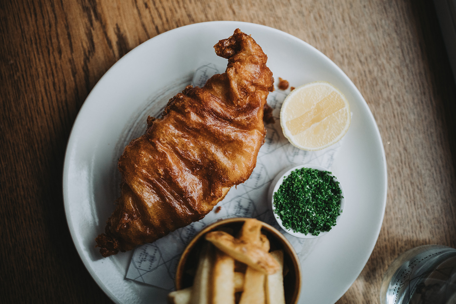 Fish and chips at the Kirkmichael Arms, a golden battered fillet on a white plate with a lemon half, a pot of chopped herbs and a cup of thick-cut chips.