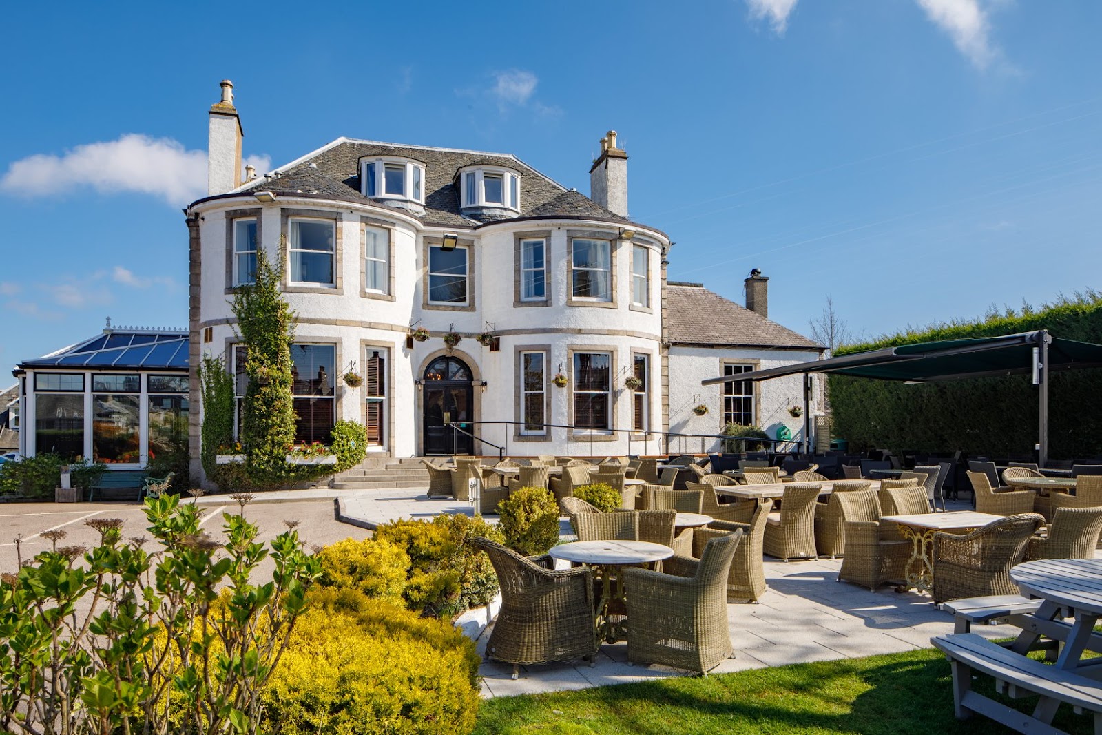 The Ferryhill House Hotel on a sunny day: a white house of 1900 with bay windows and a conservatory, and a terrace of wicker chairs and tables in front.