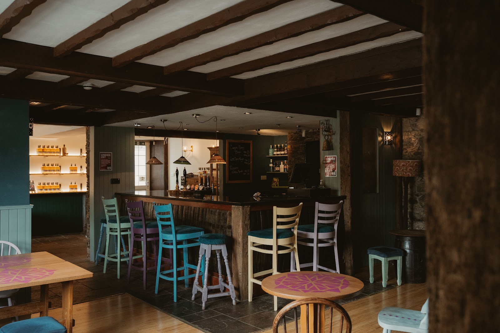 The bar of the Fintry Inn under dark ceiling beams, with painted bar stools along the counter and a lit shelf of whisky bottles in the room beyond.