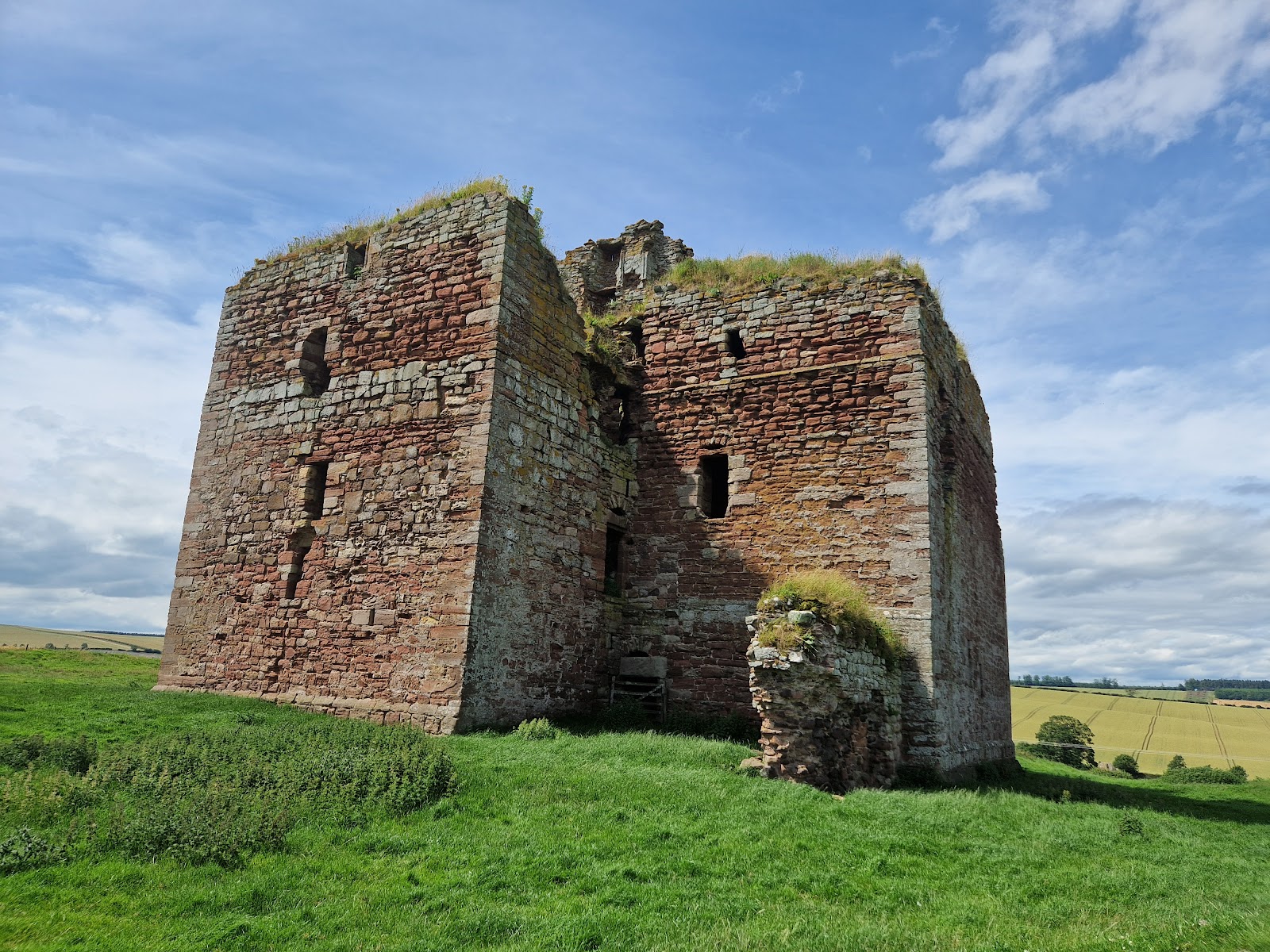 Cessford Castle, a massive roofless tower of red and grey stone, standing in green grass with farmland beyond under a blue sky.