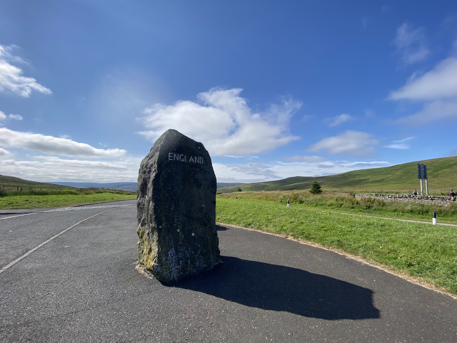 The England border stone at Carter Bar on its tarmac lay-by, green hills behind under a blue summer sky.