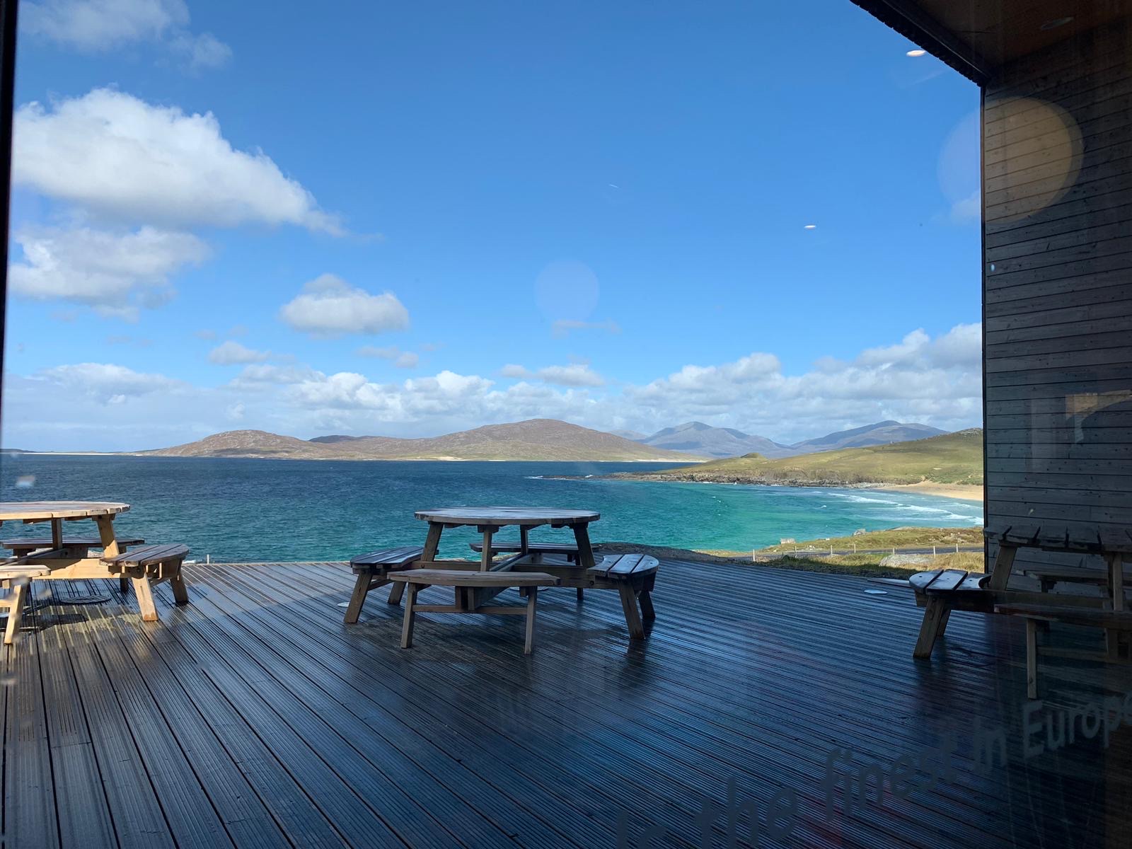 The deck at Talla na Mara, picnic benches looking out over turquoise water and white sand to the hills of Harris under a blue sky.