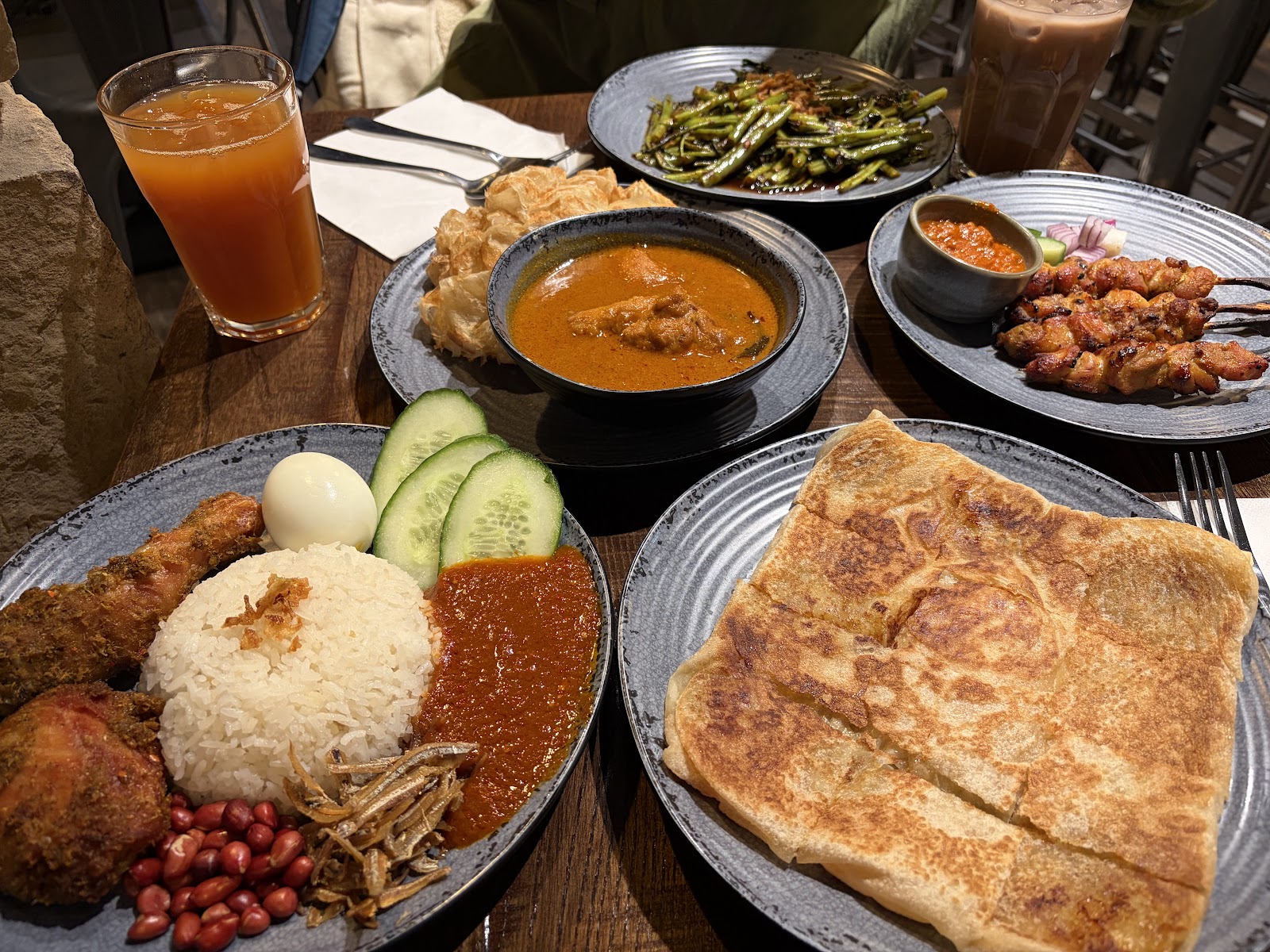 A table at Roti King: roti canai with a bowl of curry, nasi lemak with fried chicken and sambal, chicken satay, morning glory and glasses of iced tea.