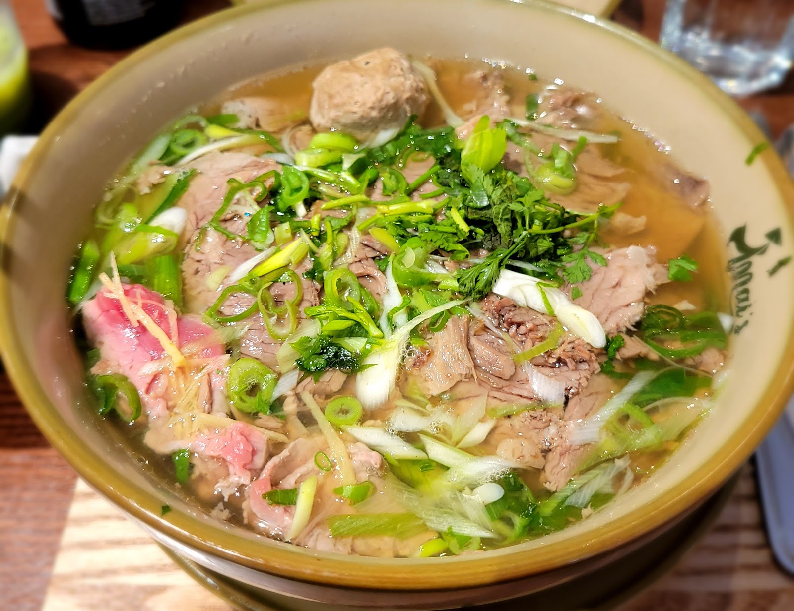 A bowl of beef pho at Mai's Vietnamese Kitchen, with sliced beef, a beef ball, spring onions and herbs in clear broth, the restaurant's name on the bowl.