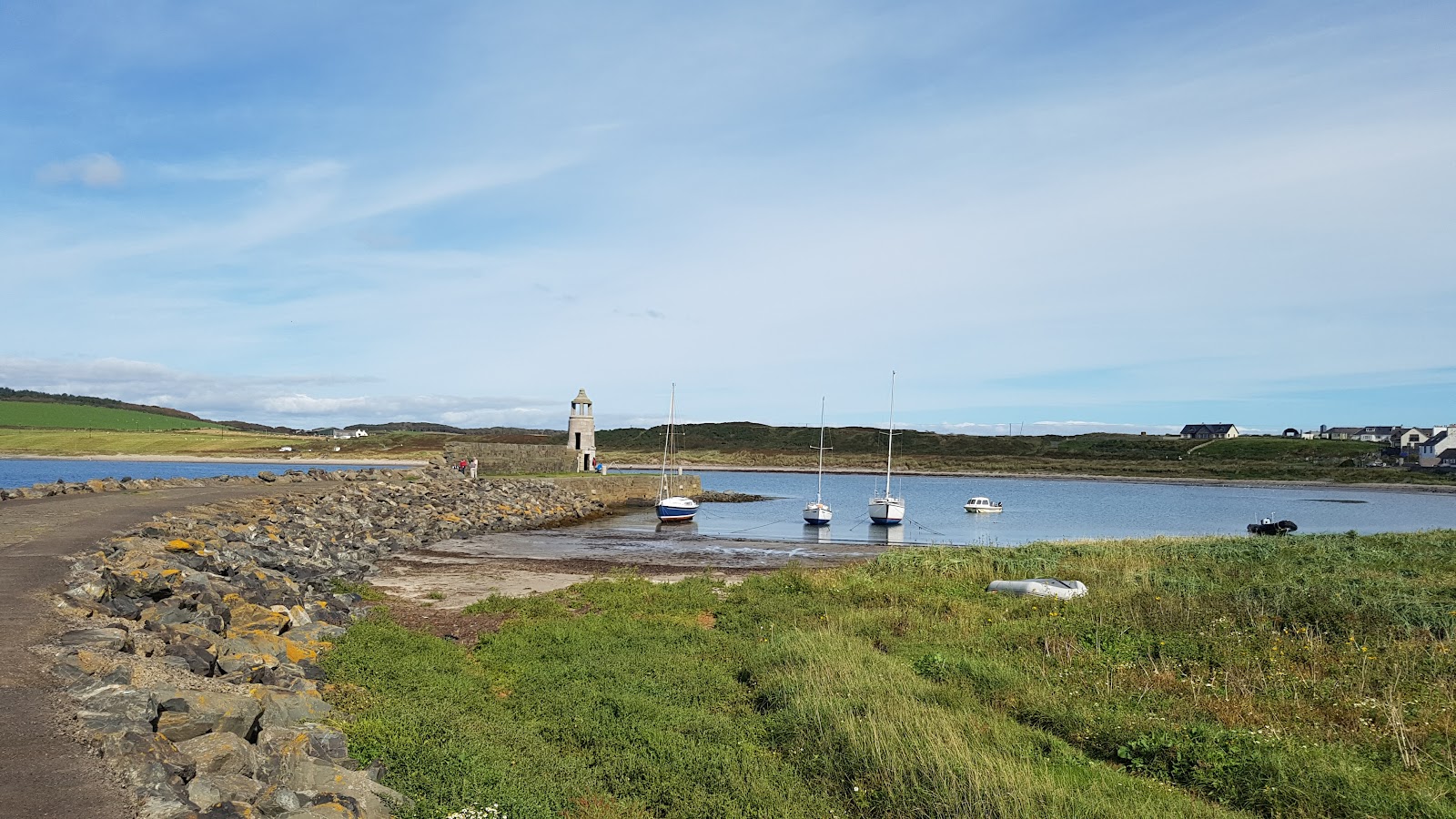 Port Logan's stone pier running out to its little round lighthouse tower, yachts moored in the calm bay and the village's houses on the far shore.