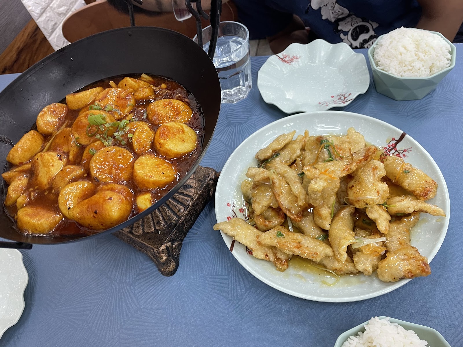 A cast-iron pot of braised tofu in a red chilli sauce beside a plate of crisp battered slices in a light, sweet sauce, on a blue tablecloth at The Way Taste.