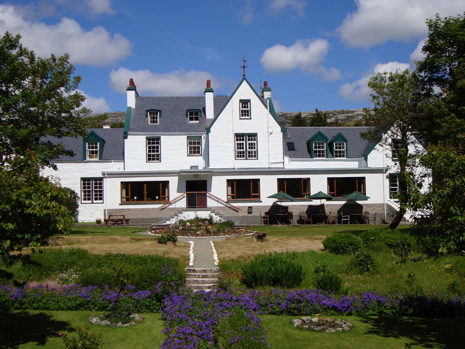 The Harris Hotel in Tarbert, a long white building with gables and dormers, above a garden of purple flowers under a blue sky.