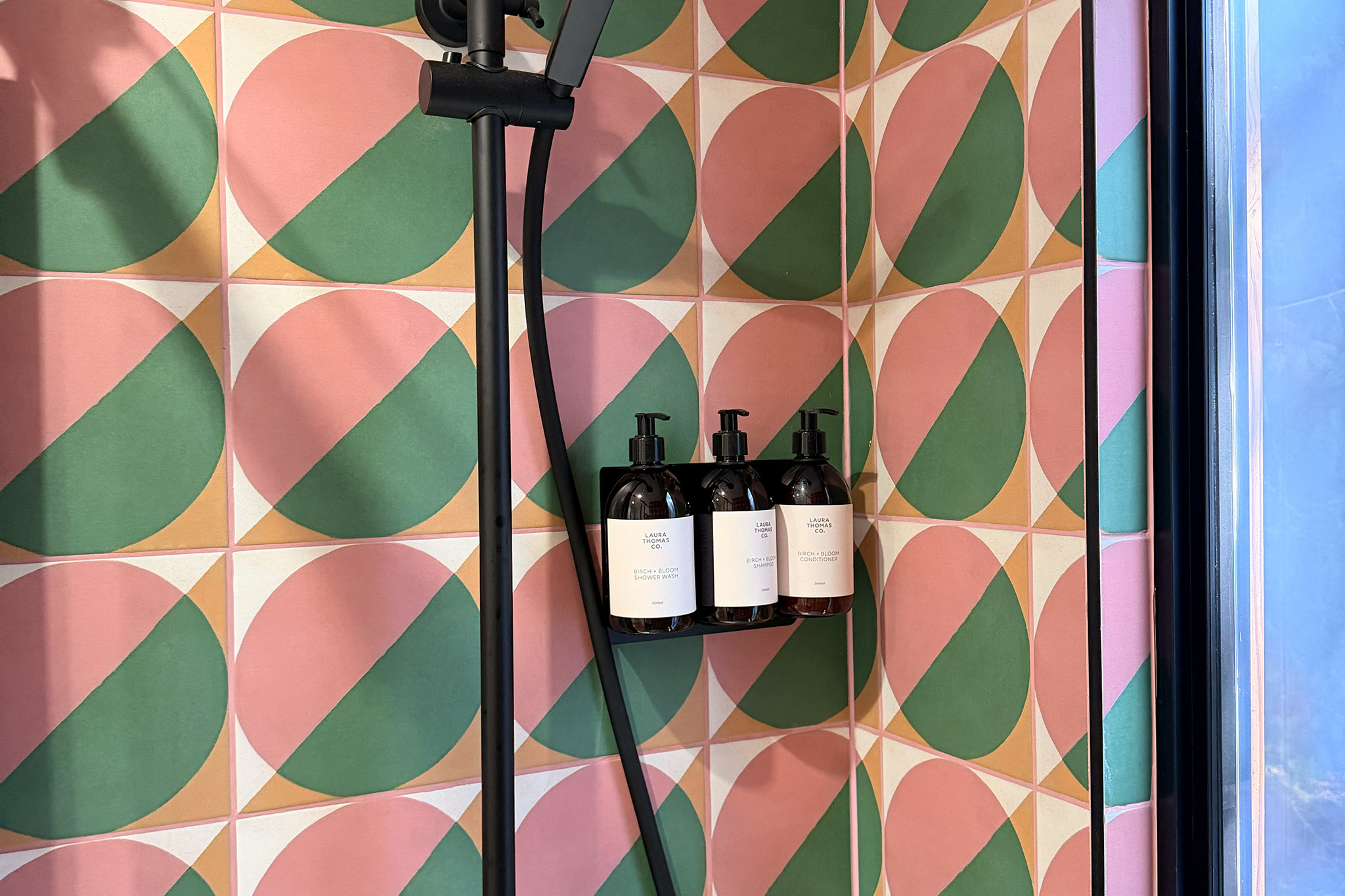 Pink and green tiles in a pattern of half-moons behind Kin's shower, three amber bottles on the wall.