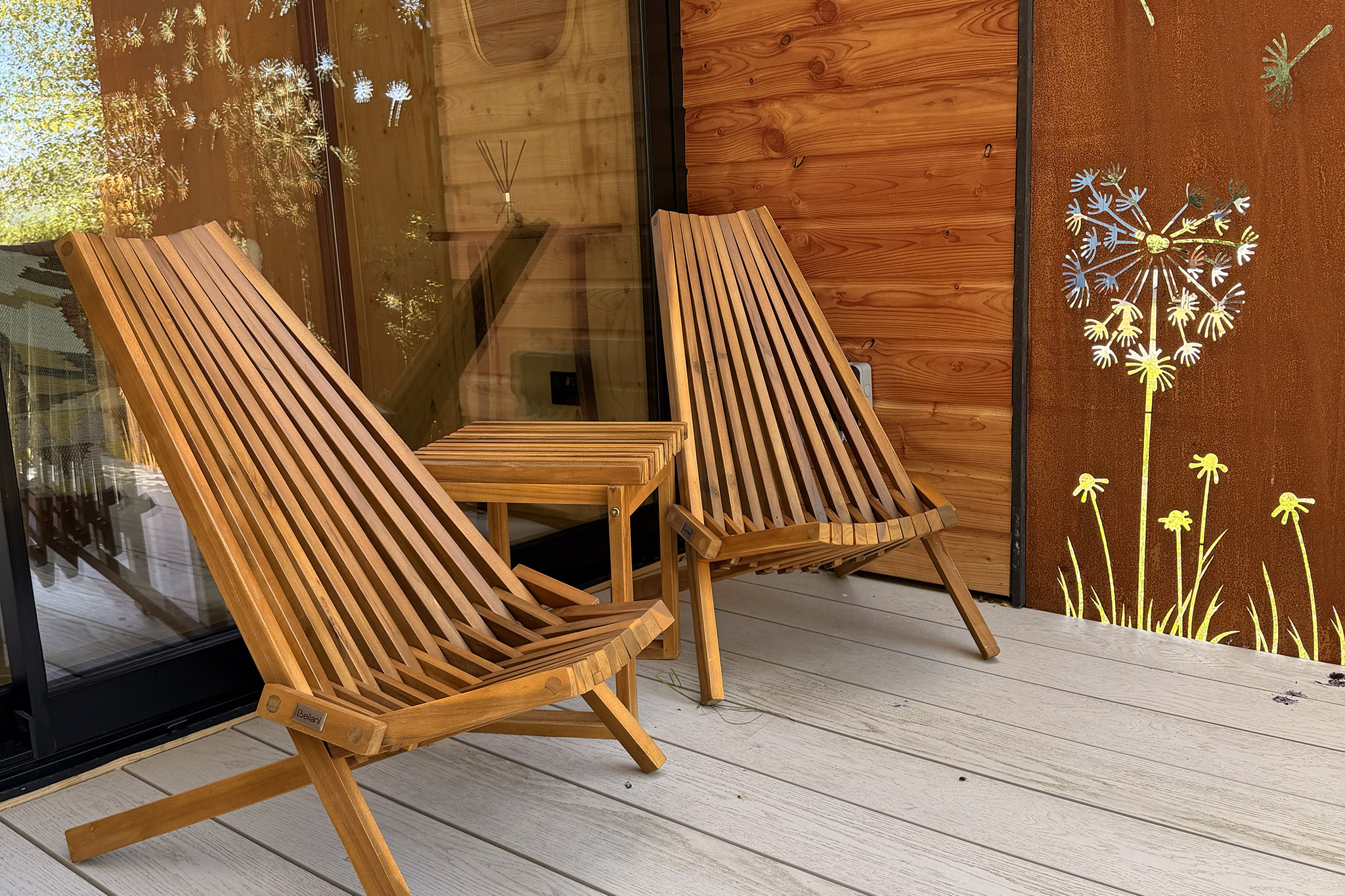 Two slatted chairs and a small table on Kin's deck, a rusted screen cut with dandelion clocks beside them.