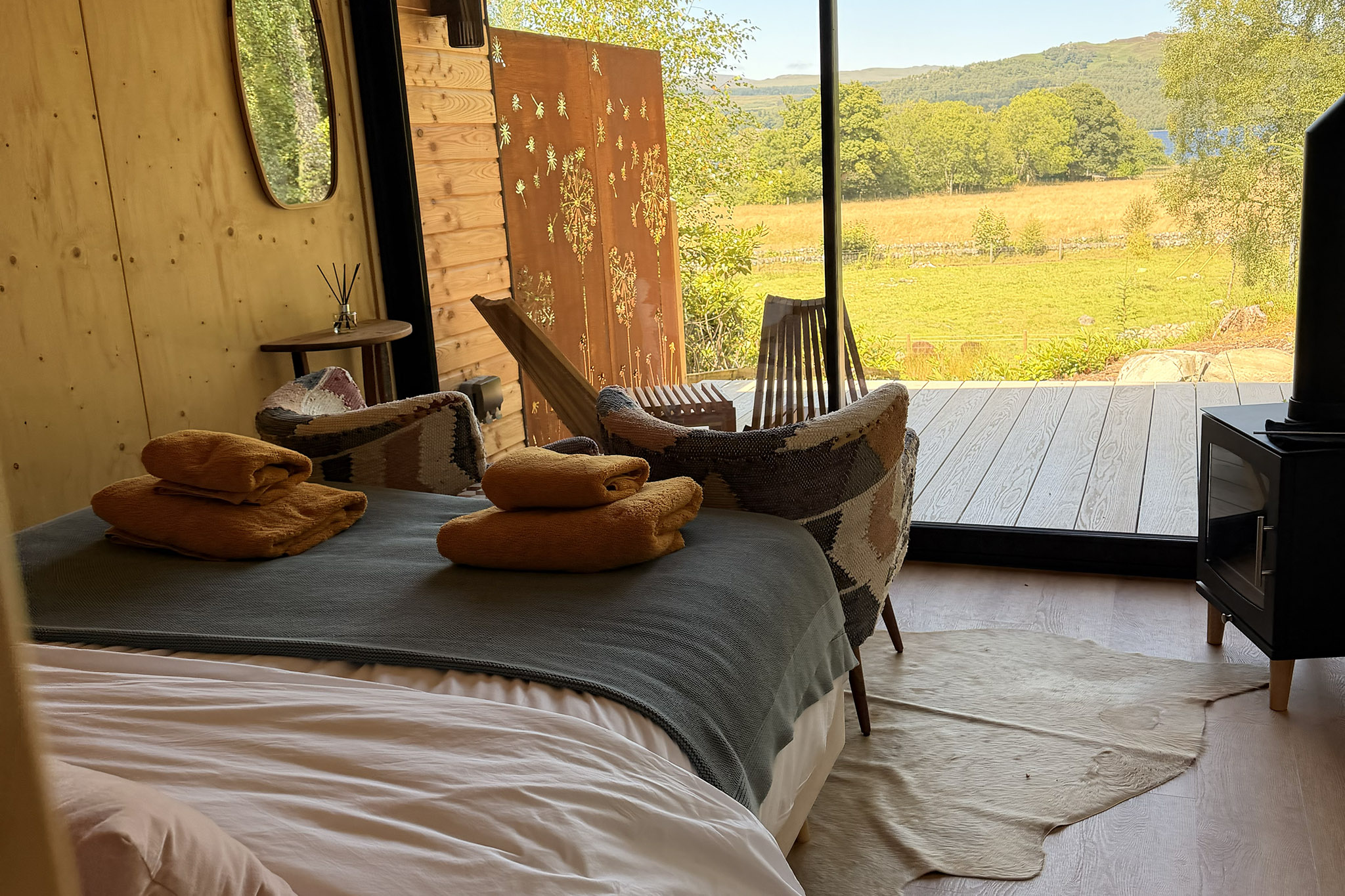 Kin's bed with orange towels and a grey throw, patterned armchairs and the deck and fields through the glass.
