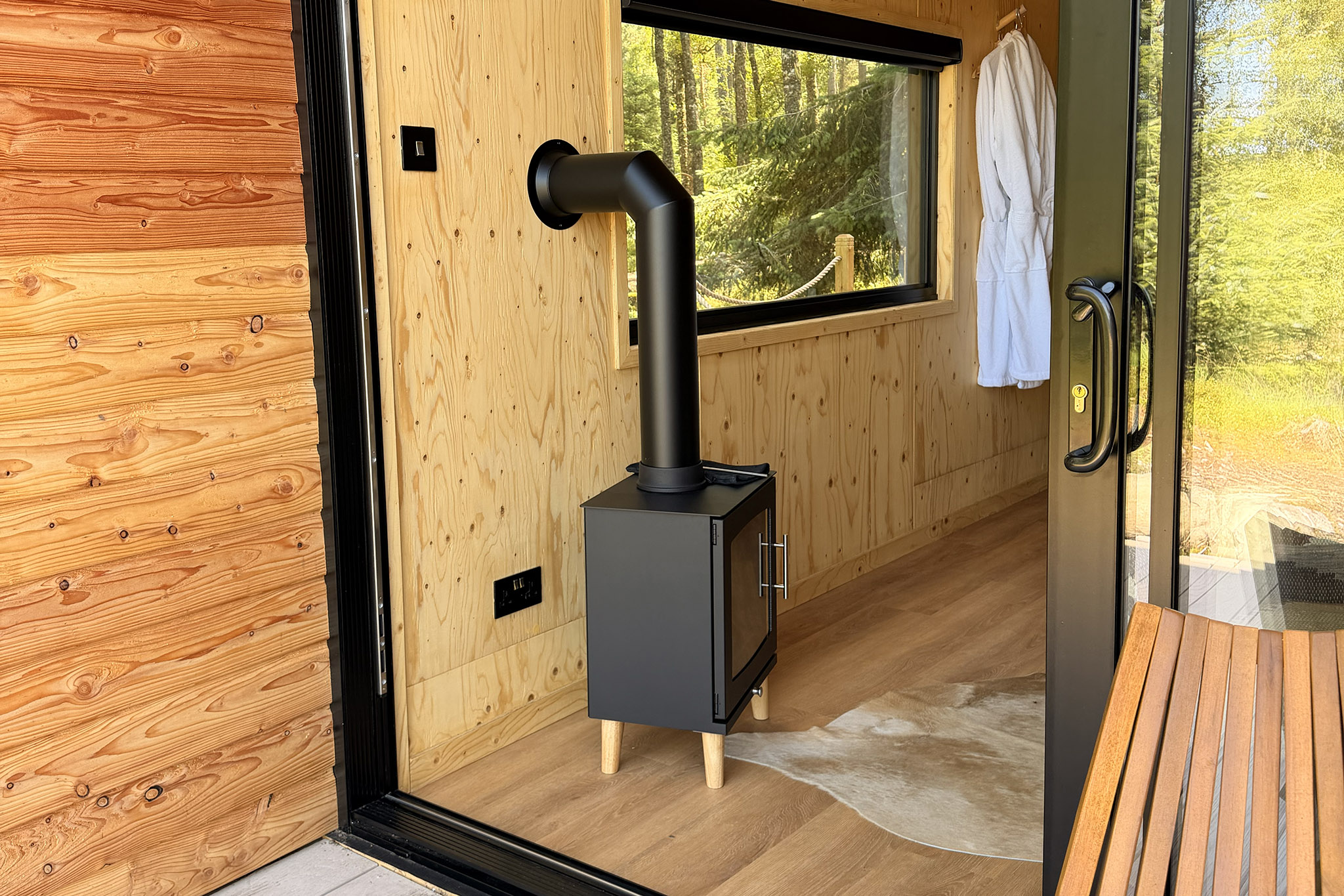 A small black stove by Kin's glass door, a white robe on its hook and the woods through the window.
