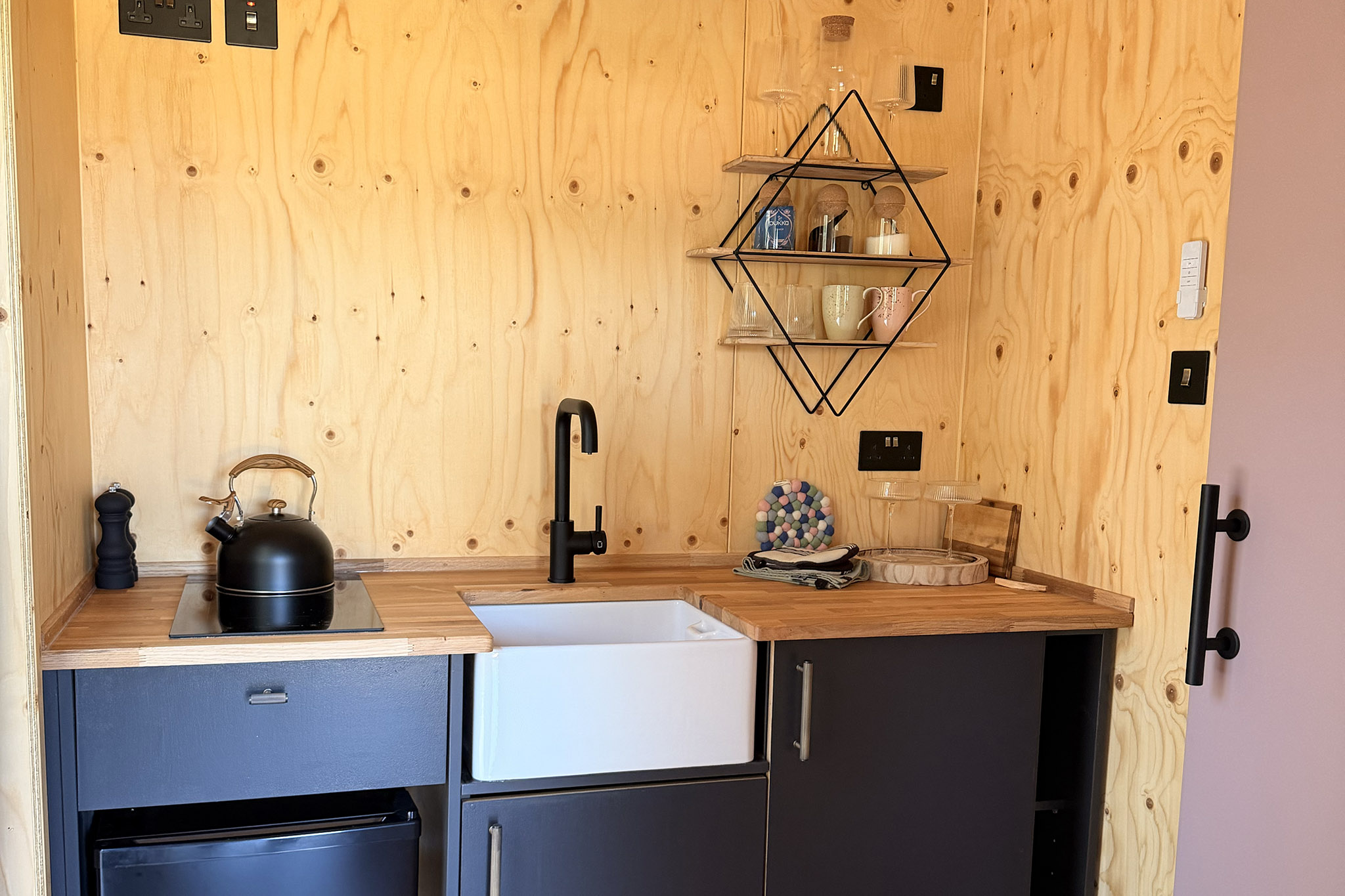 Kin's kitchenette: a Belfast sink under a black tap, a kettle on the worktop and a wire shelf of jars and cups.