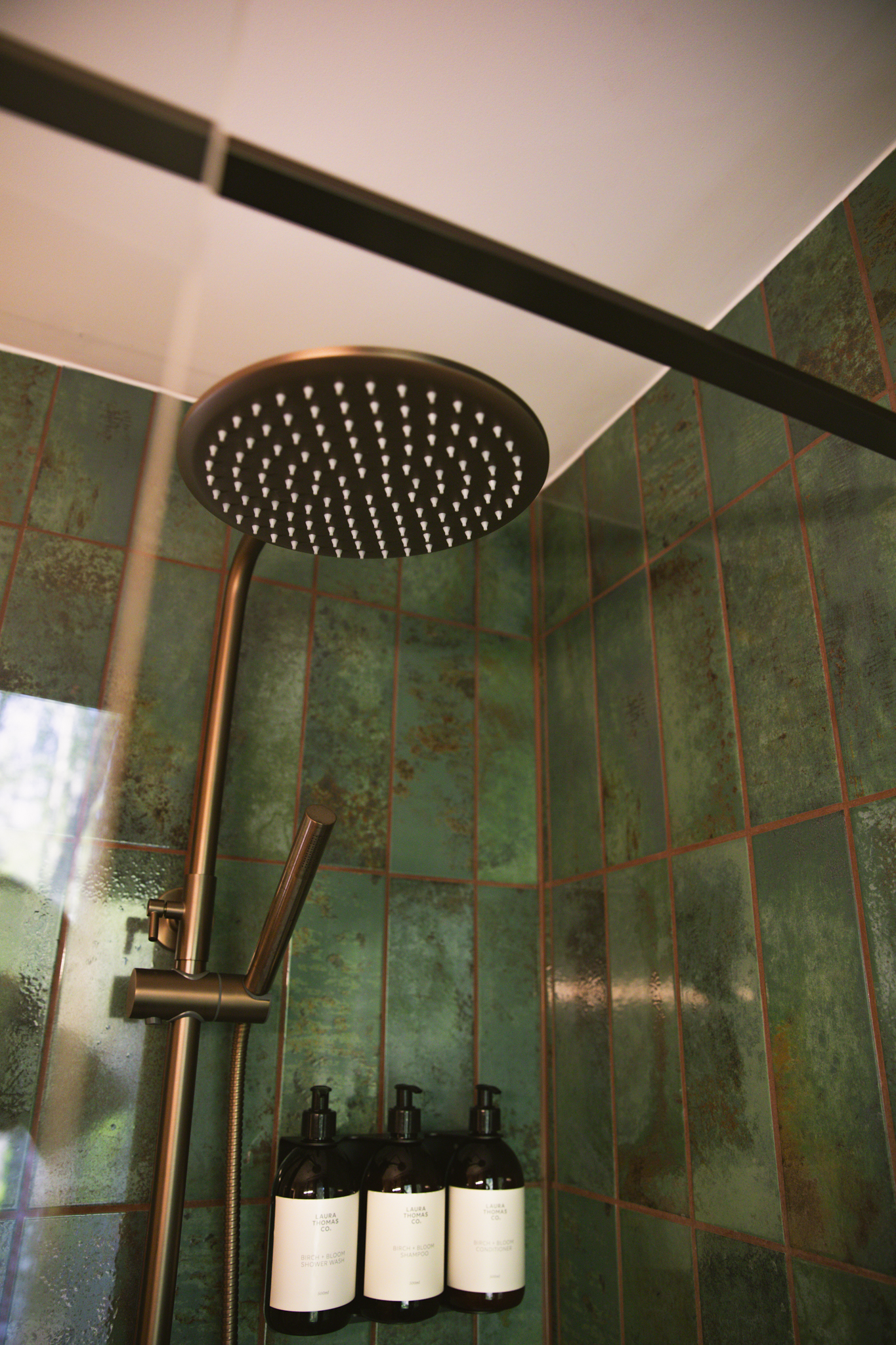A rain shower in a stall of long green tiles, three amber bottles on the wall.