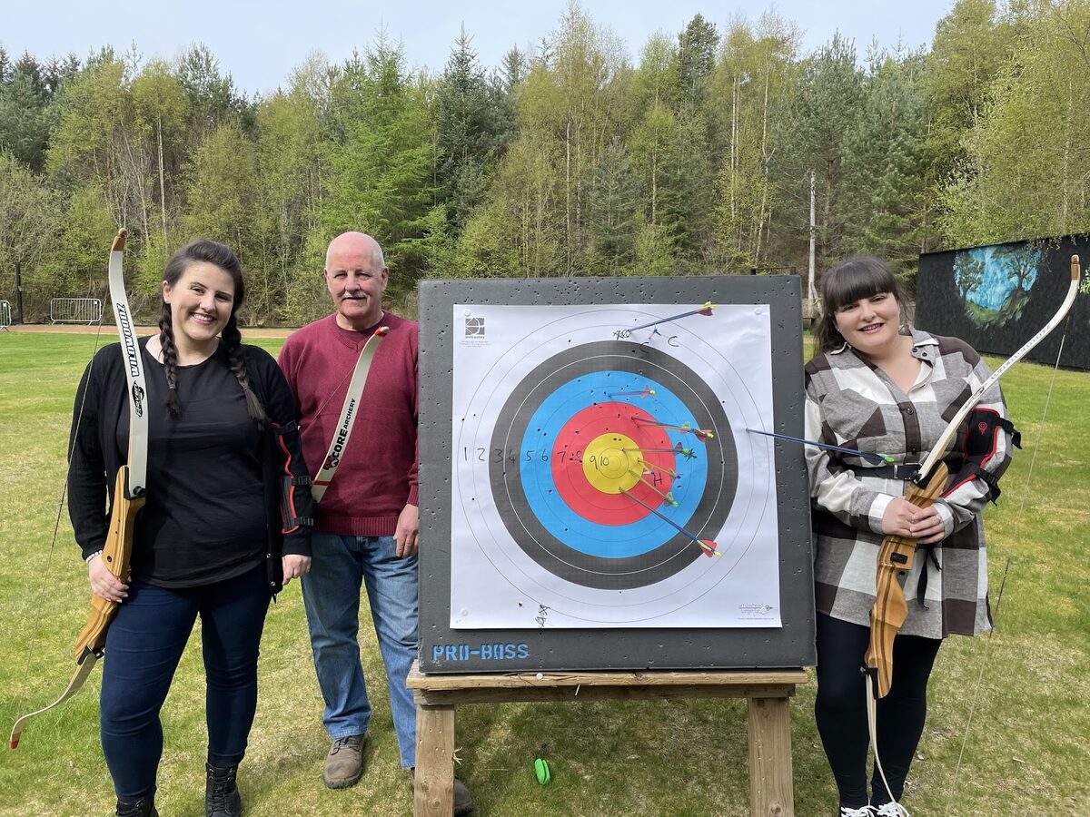 Three archers with recurve bows stand beside a Pro-Boss target scored with arrows