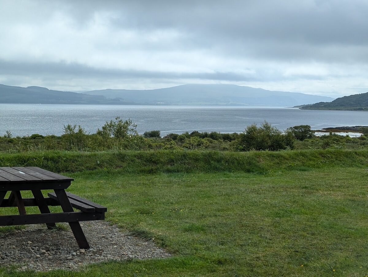 A picnic bench on the grass above Salen Bay, the calm water and hills beyond.