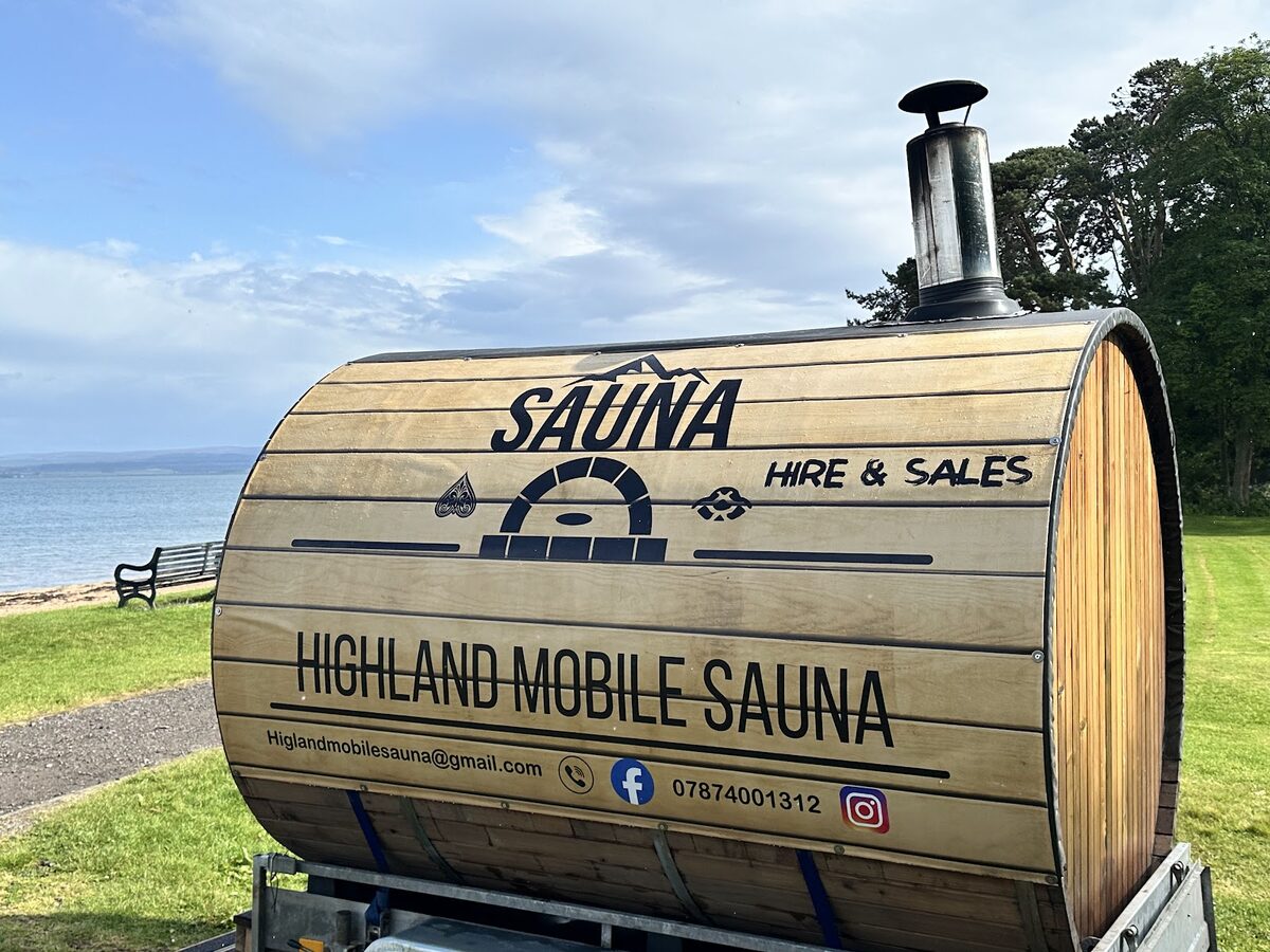 The Highland Mobile Sauna, a wooden barrel sauna on a trailer with its chimney, on the grass above Rosemarkie beach.