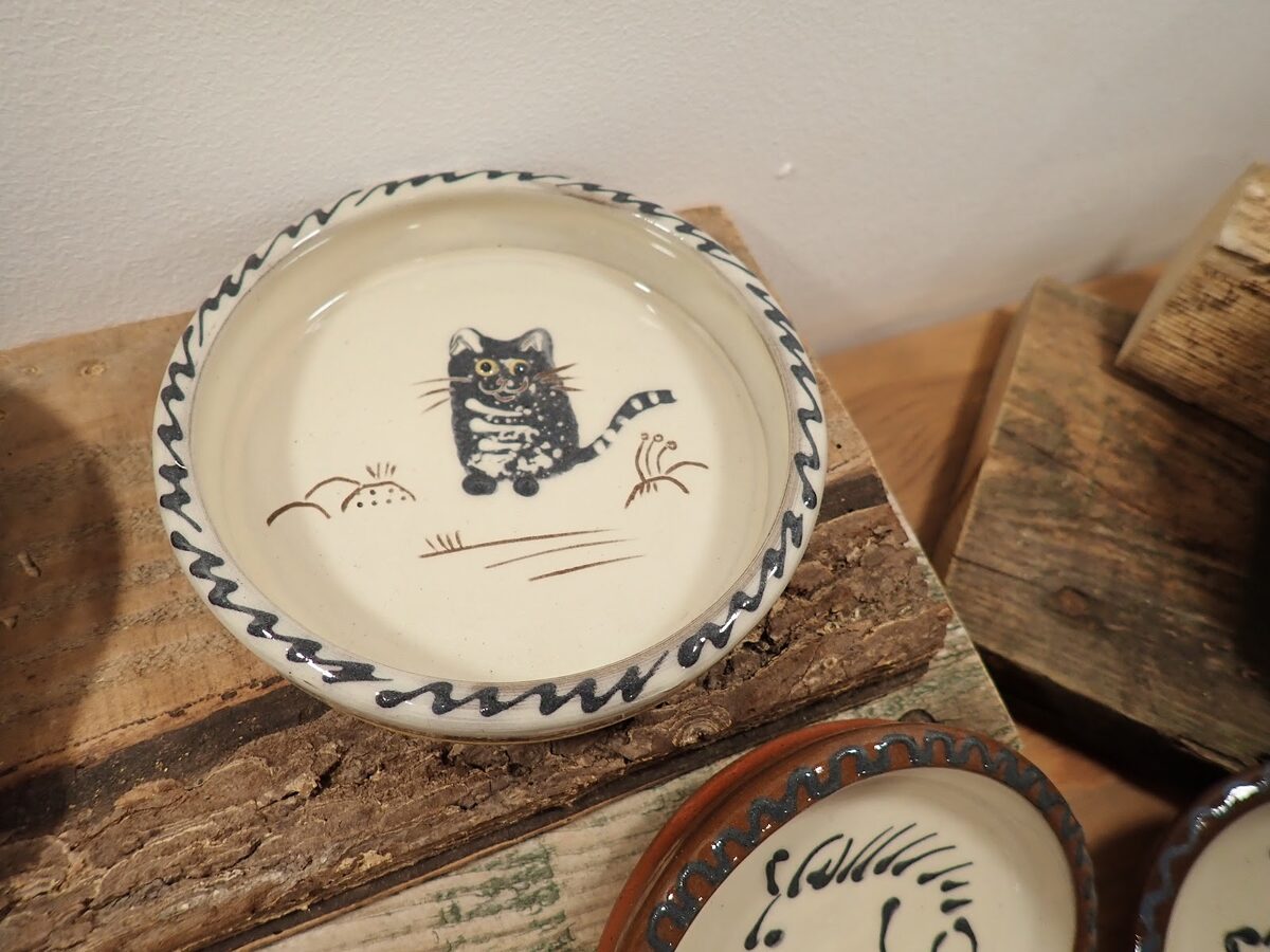 A hand-painted Cromarty Pottery dish with a striped cat, on a wooden shelf.