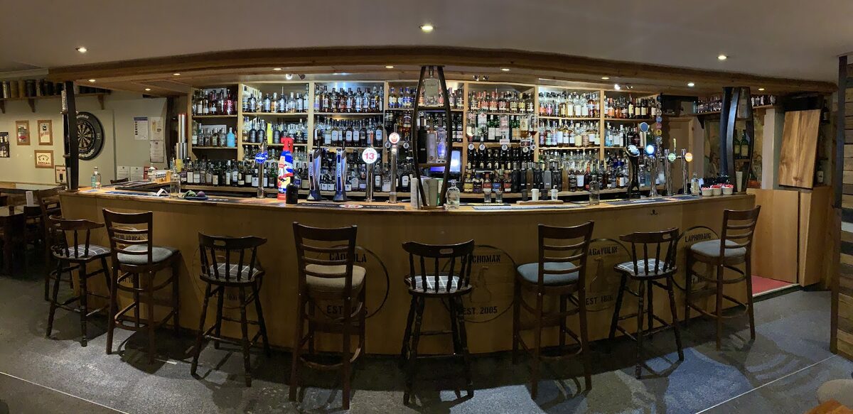 The long bar at the Ballygrant Inn, stools along the counter and shelves of whisky behind.