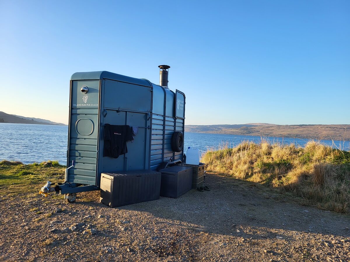 The Island Sauna Shack, a blue converted horsebox with a stove chimney, on a rocky shore by the sea.