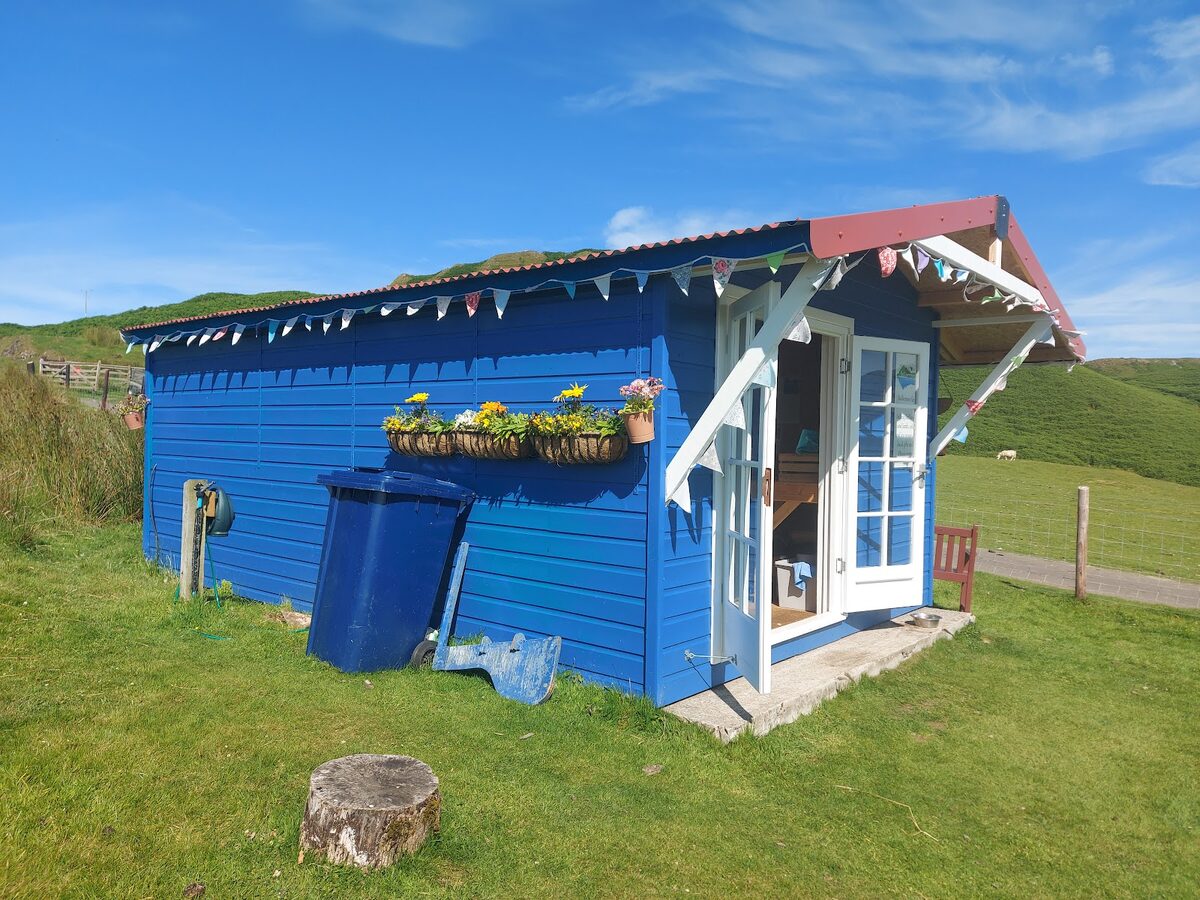 Blue wooden hut with bunting, flower baskets and open door, hills behind