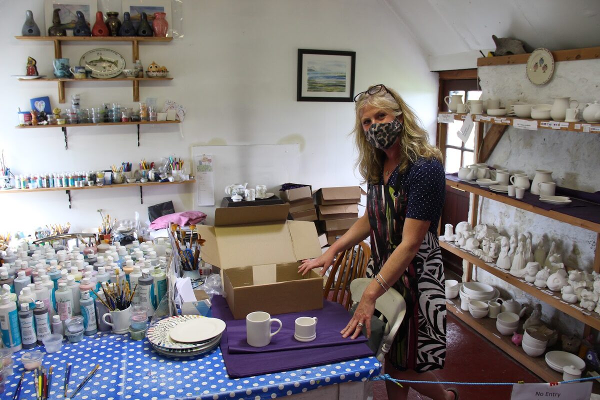 Masked woman packs boxes in a pottery studio lined with unglazed ceramics and paint bottles