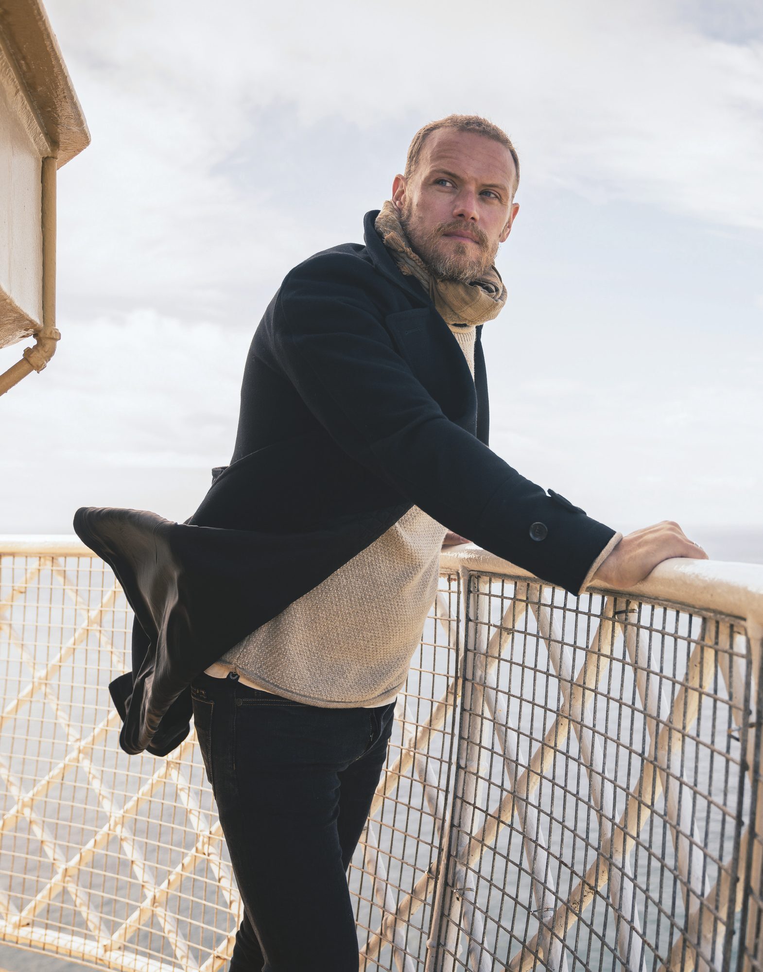 Sam Heughan leaning on a railing with sea view, opening portrait