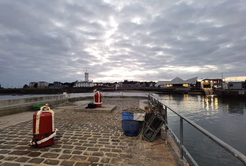 Red bollards and creels on a cobbled harbour pier, lighthouse in the distance