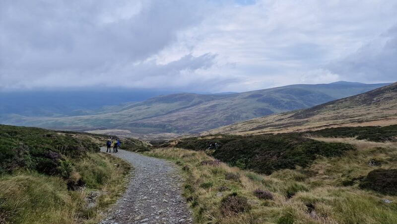 A gravel track through heather with far hills under cloud, two walkers small in the distance.