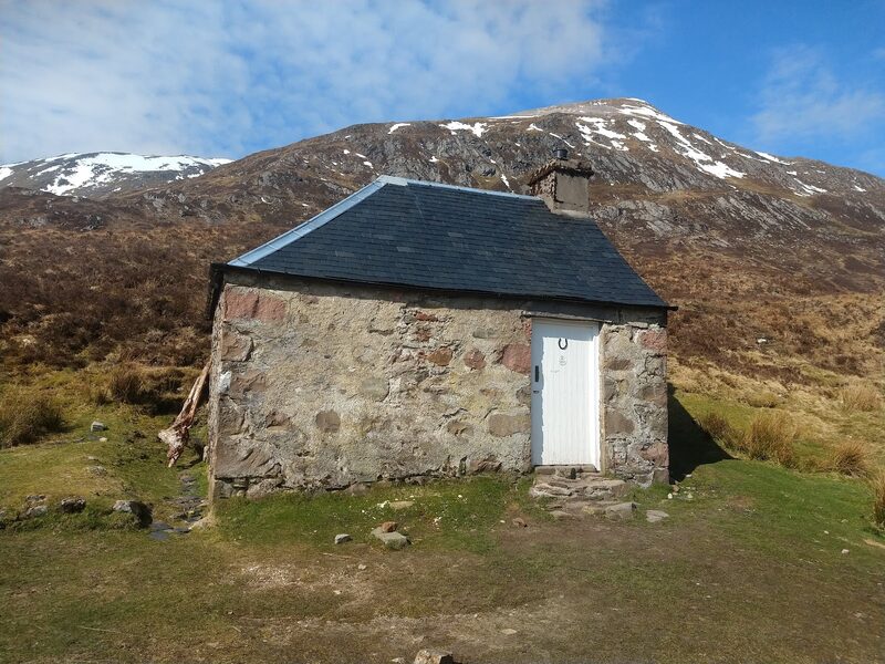 The Lairig Leacach bothy close to, rough stone walls, a white door and a slate roof, with a crag-banded hillside streaked with snow rising behind.