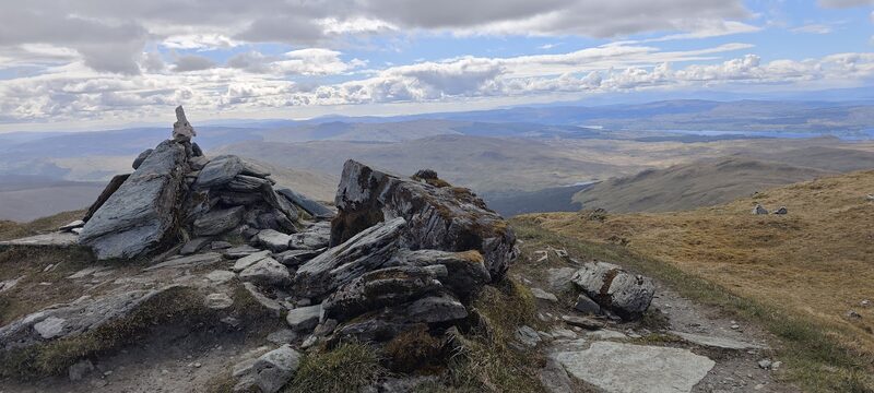 Slabs of rock and a small cairn on a hilltop, with low country and a distant stretch of water beyond.
