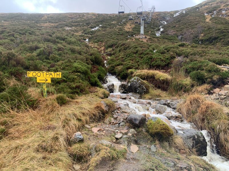 The path beside a tumbling burn under the chairlift's pylons and chairs, with a yellow sign reading Footpath.