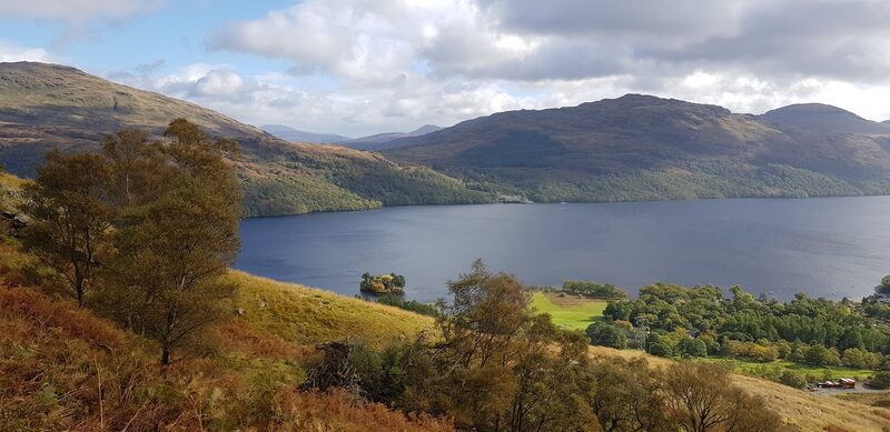 A long loch between wooded hills, seen from a bracken slope above the fields at its shore.